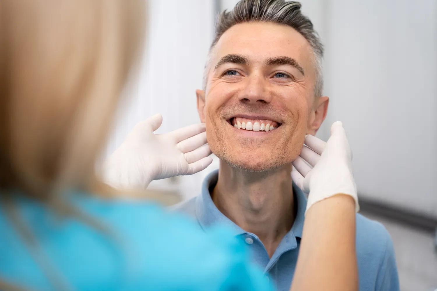 A healthcare professional examines a patient’s neck, wearing gloves, in a clinical setting. The patient is in a light blue shirt.