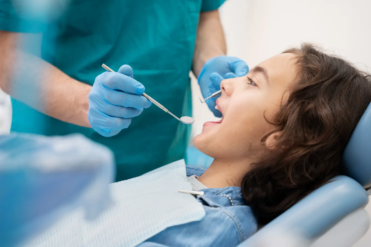 contact banner A dentist in teal scrubs and blue gloves holds a dental instrument near a patient's mouth, who is reclining in a dental chair.